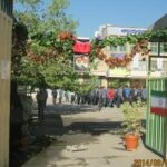 حیاط مدرسه با گل‌های تزیینی سرخ و سبز، ساختمان‌های مدرسه در اطراف، و دانش‌آموزانی که در صف یا گروهی جمع شده‌اند.