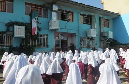 نمای خارجی ساختمان آبی مدرسه با دانش‌آموزان دختر با یونیفرم سفید و جلیقه مارون ایستاده در حیاط