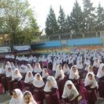 تجمع دانش‌آموزان دختر با لباس مدرسه‌ای و روسری سفید در حیاط مدرسه برای شرکت در مراسم