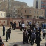 نمایی از حیاط مدرسه با دانش‌آموزانی که در حال تجمع و ایستادن در حیاط هستند، ساختمان‌های مدرسه و مناطق ورزشی در پس‌زمینه