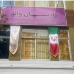 ورودی اصلی ساختمان مدرسه با تابلوی بنفش دائمی، درب‌های شیشه‌ای و پرچم‌های ملی ایران