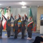 گروهی از دانش‌آموزان دختر در لباس فرم مدرسه با سریطین قرمز و طلایی که دستان خود را بالا برده‌اند در حین مراسمی جشن‌وارهٔ مدرسه با پرچم ایران در پس‌زمینه