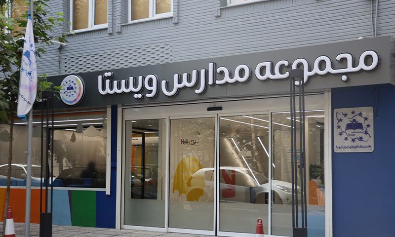 نمای بیرونی درب ورودی مدرسه با تابلوی نام دائمی و لوگو، معماری ساختمان مدرن شامل پنجره‌ها و درهای شیشه‌ای