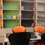 کتابخانه مدرسه با قفسه‌های رنگی (نارنجی، سبز، خاکستری، قرمز)، کتاب‌های متنوع، و صندلی‌های نارنجی و سیاه برای مطالعه