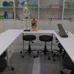 نمای داخلی آزمایشگاه علوم مدرسه با میز سفید، صندلی‌های سیاه، کابینت‌های ذخیره‌سازی، و مدل اسکلت انسانی