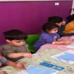 گروهی از دانش‌آموزان در حال انجام فعالیت نقاشی و هنری با مواد و رنگ‌های مختلف در کلاس درس با دیوار صورتی