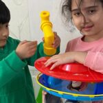 دو دانش‌آموز کوچک در حال بازی با اسباب‌بازی‌های رنگی شامل هولا‌هوپ و دیگر اسباب‌بازی‌های پلاستیکی در محیط بسته
