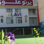 نمای بیرونی ساختمان مدرسه علوی جنت‌آباد با تابلوی نام و تزیینات کودکانه، گلهای بنفش در پیشنمایش