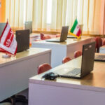 کلاس کامپیوتری مجهز به میزهای دسته‌ای با لپ‌تاپ و کامپیوتر رومیزی، صندلی‌های قرمز و تابلو سفید
