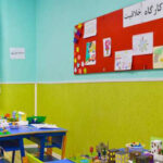کلاس درسی برای فعالیت‌های خلاقیت و هنر با میز کار رنگی، تجهیزات، و نقاشی‌های دانش‌آموزان بر روی دیوار قرمز