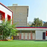 نمای خارجی ساختمان مدرسه علوی منیریه با معماری مدرن، نوارهای قرمز و سفید، پنجره‌های شیشه‌ای، و حیاط سبز روبروی ورودی