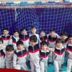 گروهی از دانش‌آموزان پسر با لباس ورزشی قرمز و سفید در سالن ورزشی، پشت دروازه هندبال برای عکس تیمی