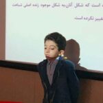 دانش‌آموز پسر با لباس مدرسه در حال ارائه شفاهی جلوی صفحه‌نمایش‌ای که متن فارسی درس را نشان می‌دهد