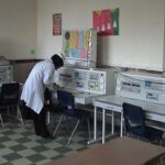 نمای سایت کامپیوتر مدرسه با چندین ایستگاه کاری، مانیتورهای رایانه‌ای، صندلی‌های مشکی، و فردی در حال بررسی و کار با تجهیزات