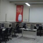 نمایی از سایت کامپیوتری مدرسه با چند ایستگاه کار مجهز به رایانه‌های رومیزی، صندلی‌های اداری و تخته سفید