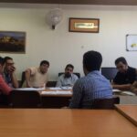 نشست پرسنل و معلمین در کلاس درس؛ گروهی از مردان دور یک میز نشسته در حال برگزاری جلسه رسمی با تابلوی سفید و نقاشیات دیواری در پس‌زمینه