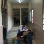 دانش‌آموز نشسته بر روی صندلی آبی در راهروی مدرسه در حال خواندن و نوشتن، با درهای کلاس‌های درسی در پس‌زمینه
