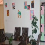 فضای داخل کلاس درس با صندلی‌های پشتی مشکی، میز، تابلوهای هنری رنگی روی دیوار، گیاهان تزیینی و حمام‌های تغییر لباس رنگی در پس‌زمینه