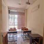 نمای داخلی کلاس درس مدرسه با میزهای تحصیلی و صندلی‌های قهوه‌ای، پنجره‌های بزرگ و تجهیزات آموزشی