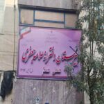 تابلوی دائمی ورودی مدرسه با متن فارسی، بناهای اطراف و سبزیجات آتش‌خودری