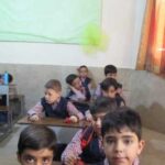 دانش‌آموزان در یک کلاس‌درس نشسته در پشت میز و صندلی، پرده نارنجی در پس‌زمینه، تخته سبز روی دیوار