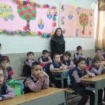 دانش‌آموزان در کلاسی نشسته بر پشت میزهایشان در حال انجام فعالیت درسی و معلم در پس‌زمینه