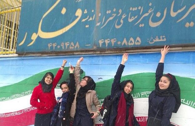 گروهی از دانش‌آموزان دختر ایستاده در مقابل تابلوی آبی مدرسه با دستان بالا در حین مراسمی گروهی
