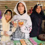 تصویری از یک رویداد خیریه مدرسه‌ای با دانش‌آموزان دختری دور یک میز پر از غذاهای خانگی و شیرینی‌ها در حال فروش به‌منظور جمع‌آوری کمک‌های خیریه