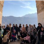 گروهی از دانش‌آموزان در یک سایت باستان‌شناسی با دیوارهای سنگی باستانی و کوه‌های پس‌زمینه، حین فعالیت بازدید میدانی