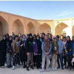 دسته‌ی عکس یادگاری دانش‌آموزان ایستاده در حیاط مدرسه به سبک معماری اسلامی با آرچ‌های قوسی و دیوارهای خشتی
