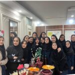 گروهی از دانش‌آموزان دختر در حین مراسم جشن و جمع‌آوری در فضای داخلی مدرسه، با میز پر از خوراکی‌های مختلف و کیک‌های تزیین‌شده