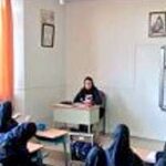 کلاس درس با دانش‌آموزان نشسته و معلم ایستاده در جلو، تابلوی کلاس و پنجره‌های روشن در پس‌زمینه