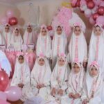 گروهی از دانش‌آموزان دختر با حجاب سفید در مراسمی جشن‌وار که با بالون‌های صورتی و تزئینات فرشته‌ای تزیین شده است