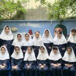 گروه‌عکسی از دانش‌آموزان دختر با لباس یکدست‌ی فرم مدرسه در حیاط، جلوی دیوال نقاشی‌شده‌ی رنگی