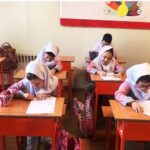 دانش‌آموزان دختری با حجاب در یک کلاس درس روی میزهای قرمز و صندلی‌های چوبی نشسته، مشغول انجام تکالیف درسی