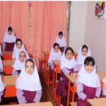گروهی از دانش‌آموزان دختر ایرانی با لباس مدرسه و حجاب در یک کلاس درس نشسته‌اند. کلاس دارای صندلی‌های نارنجی، میز‌های چوبی، دیوارهای سفید و پوسترهای آموزشی رنگی است.