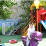 حیاط مدرسه با تجهیزات بازی رنگی برای دانش‌آموزان، دیوارنقاشی‌شده و درخت سایه‌دار