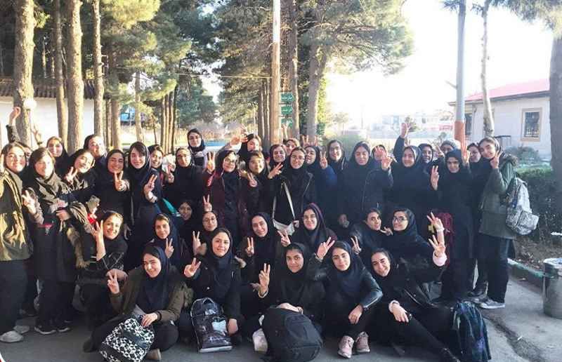 گروهی از دانش‌آموزان دختر با حجاب اسلامی در حیاط یا راهروی مدرسه‌ای کنار درختان، به مناسبت یک رویداد یا مراسم مدرسه‌ای، با دستان بر سینه و لبخند برای دوربین.