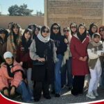 گروه دانش‌آموزان دختر و یک کودک که برای عکس یادگاری کنار هم ایستاده‌اند، در حیاط مدرسه با تابلوی اطلاعات در پس‌زمینه