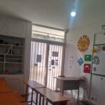 داخل یک کلاس درس با میزهای چوبی نارنجی، تخته سفید، قفسه‌های ذخیره‌سازی، و تزئینات آموزشی روی دیوارها