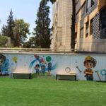 حیاط مدرسه با دیوار نقاشی‌شده با تصاویر کاراکترهای رنگین، چمن مصنوعی سبز، و ساختمان