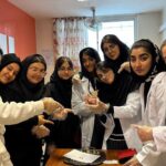 دانش‌آموزان دختر در حال انجام یک آزمایش علمی تحت نظارت معلم در کلاس درس، با دستکش سفید و مواد آزمایشی