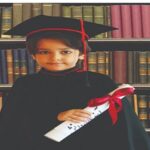 دانش‌آموزی با جبه و کلاه دکتری و دیپلم فارغ‌التحصیلی در کنار قفسه‌های کتاب
