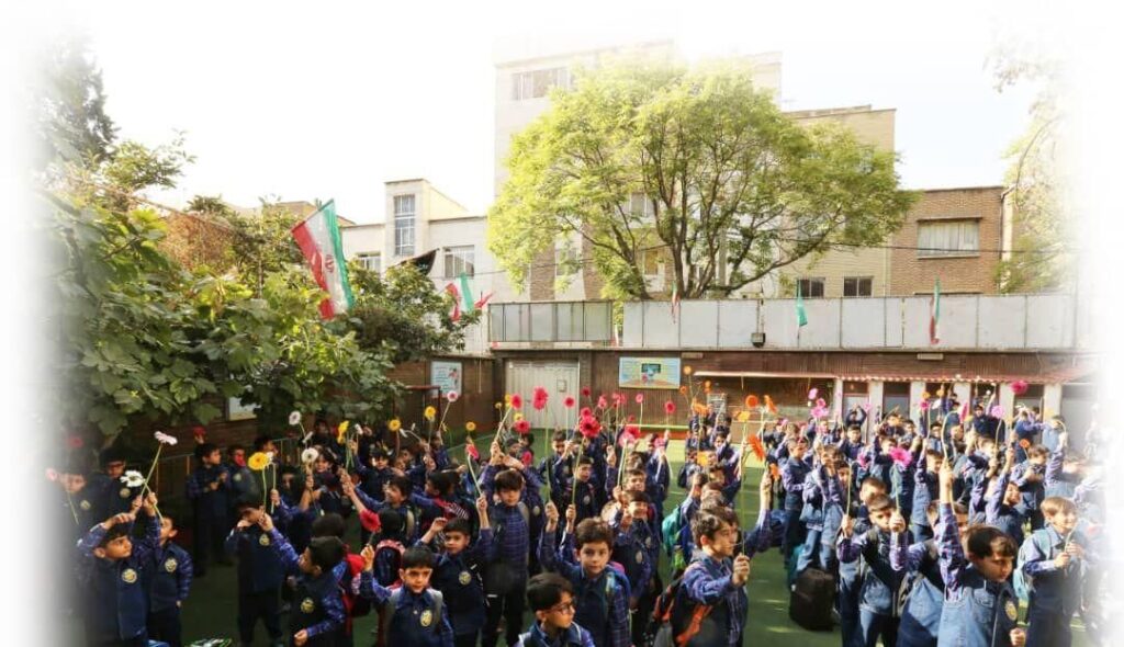 دانش‌آموزان مدرسه در حیاط با پرچم و گل به‌مناسبت جشن ملی شرکت می‌کنند