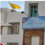 نمای بیرونی سردر و ورودی اصلی ساختمان مدرسه با تابلوی آبی، پرچم زرد، و معماری خاکی