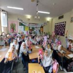 دانش‌آموزان دختر با حجاب در یک کلاس درسی روشن و مجهز به میز، صندلی، و مواد آموزشی روی دیوارها