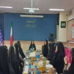 دانش‌آموزان و اساتید در حال شرکت در مراسمی گردهم‌آیی‌ای دور میز غذایی با تزئینات بادکنک‌های بنفش و سفید و پرچم ایران