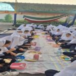 دانش‌آموزان دختری مدرسه با لباس سفید و مشکی در حال شرکت در فعالیت نقاشی جمعی در حیاط مدرسه