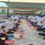 دانش‌آموزان نشسته در ردیف‌های منظم در حیاط مدرسه، هنگام برگزاری مراسمی آموزشی با پرچم‌های آیینی در پس‌زمینه