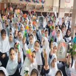 دانش‌آموزانی با حجاب سفید در حال شرکت در مراسم یادبود داخل سالن مدرسه، با دسته‌های گل رنگی و عکس‌های شهدا در دست و پس‌زمینه